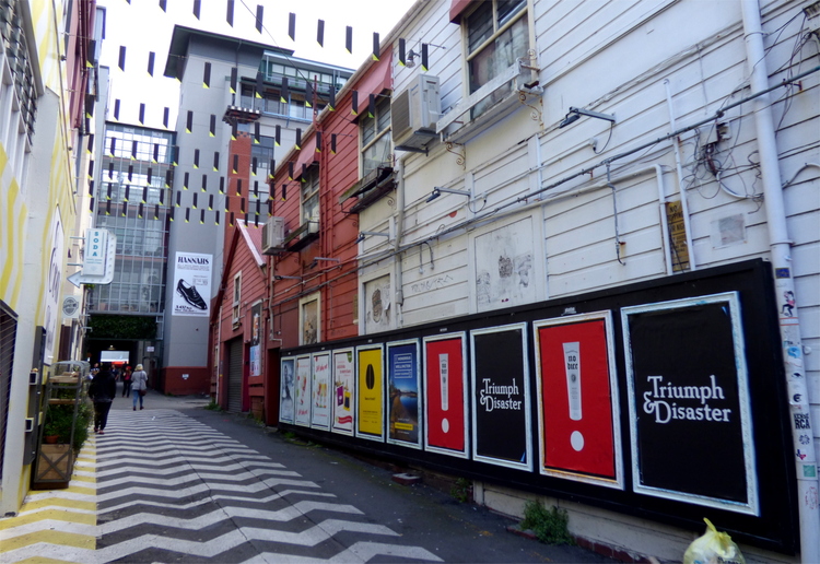 Graphic posters in a side alley, some of them showing the words 'Triumph & Disaster' in white on black