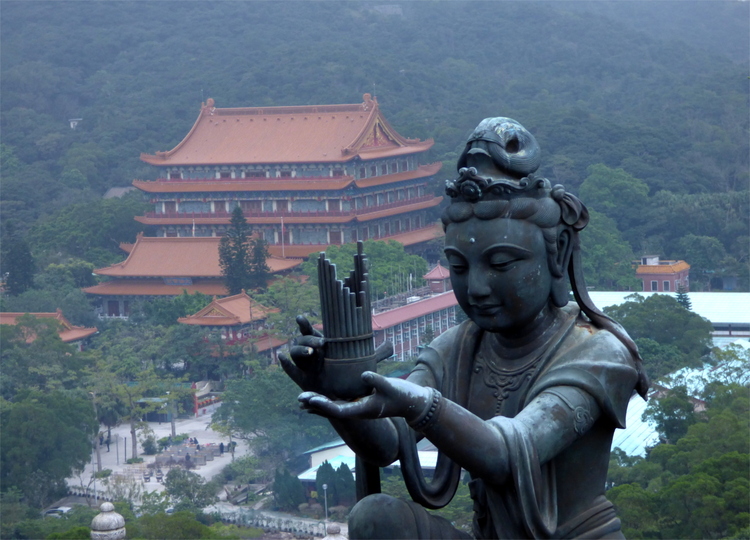 A large metal statue of a woman holding some musical instrument as an offering with a temple building in the background