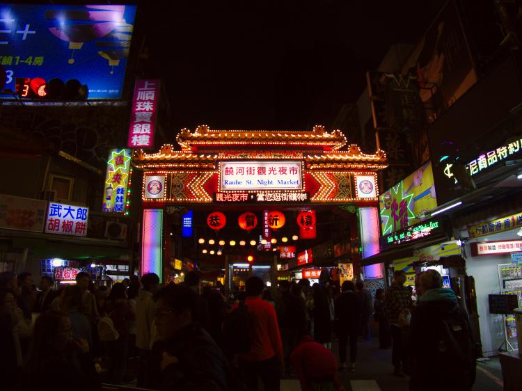 People passing though a lit-up free-standing gate to a night market