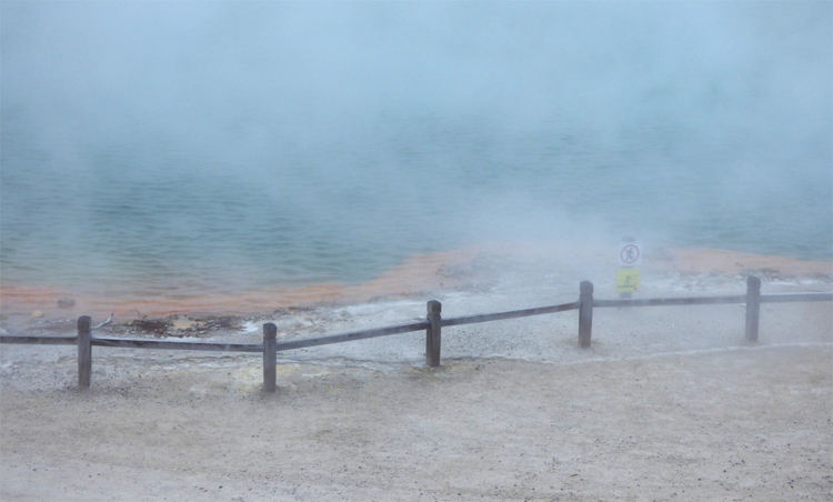 A low wooden fence around a geothermal pond visible through dense white vapor