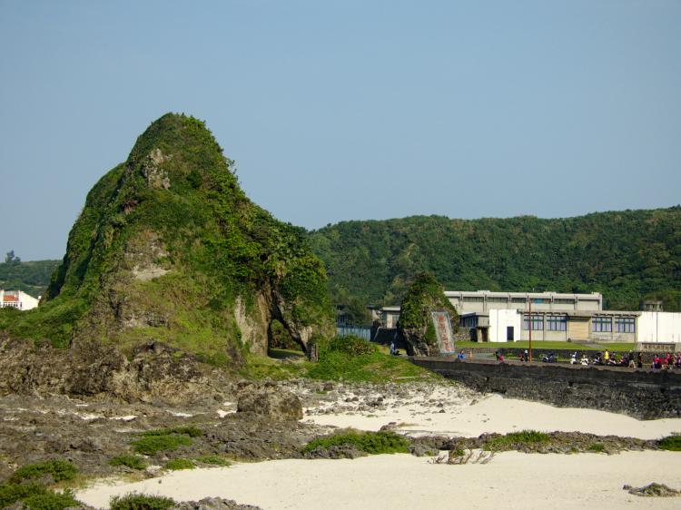 A rock formation on a beach of fine white sand with green hills in the background