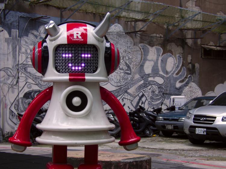 A sculpture of a red-and-white horned robot wearing large headphones with street-art in the background