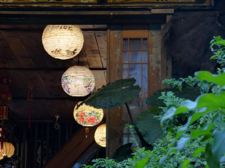 Round white paper lanterns with floral illustrations hanging from the ceiling of a wooden room viewed through an open terrace door