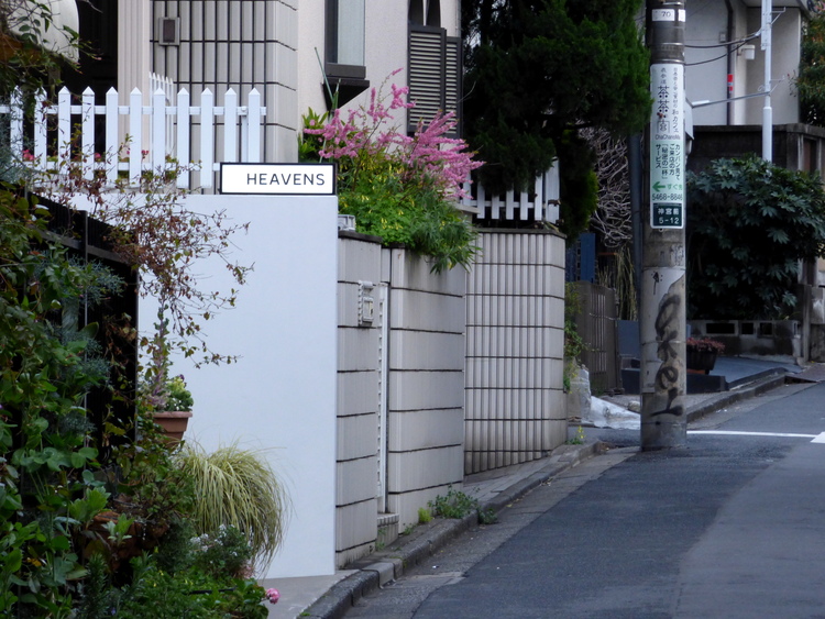 The side of a street in Tokyo with a small sign on one of the walls reading 'HEAVENS' and some flowers behind it