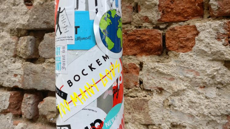 A lamppost covered in stickers in front of a rough red brick wall, one of the stickers reading 'too many gods' in a jagged yellow font