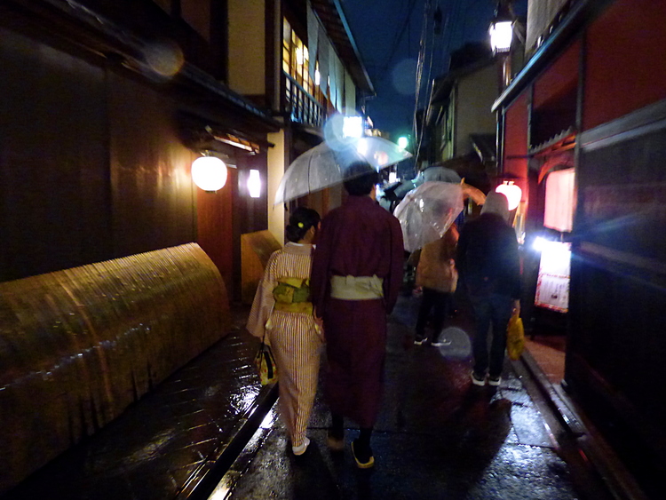 People walking through a narrow pedestrian street in the rain, among them a couple in traditional Japanese clothing, holding a large transparent umbrella