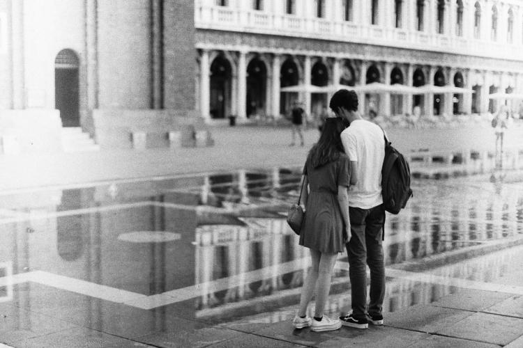 A young couple leaning against each other standing on the edge of a puddle on a public square