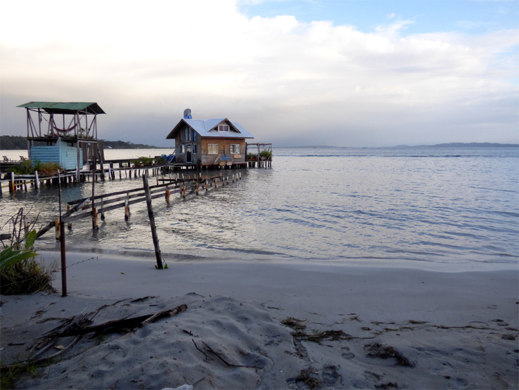 A wooden pier running out into the ocean from a beach, leading to a tiny wooden house out on the water.