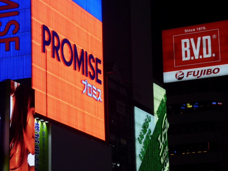 Large, lit-up advertisement screens featuring Japanese and English writing, one of them a red square reading 'PROMISE'