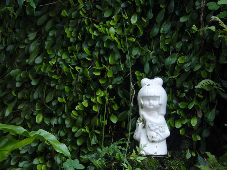 A small, white porcelain figurine of a child placed in front of a wall of greenery