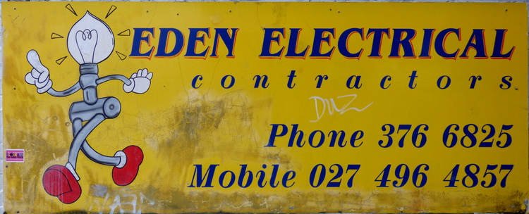 A slightly withered yellow metal sign showing a cartoon character made of wire with a lightbulb for a head reading 'Eden Electrical contractors' alongside some phone numbers