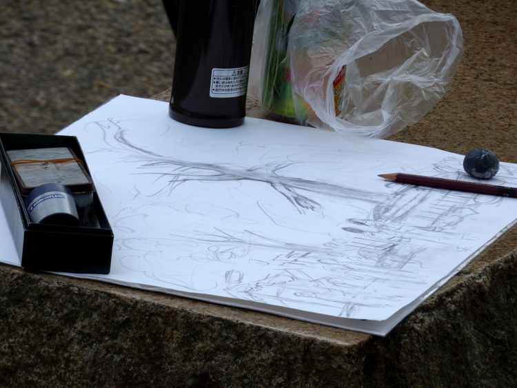 A sketchbook with a graphite sketch of a tree resting on a stone bench with pencils and other drawing utensils