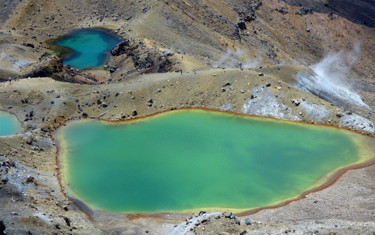 A turquoise-green volcanic pool fading to yellow and red at the edges in a barren, rocky landscape