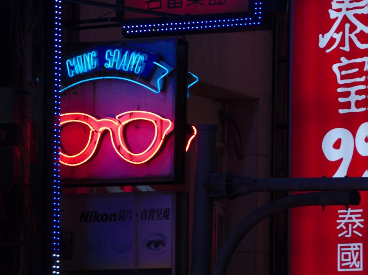 A neon sign showing stylized red glasses blue text above next to other advertising signs