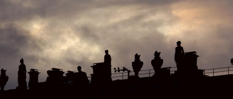 Silhouettes of different sculptures standing on top of a building against a cloudy evening sky