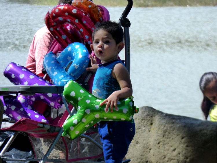 A young child standing next to a stroller holding a green Y-shaped balloon making an o-Face, with more balloons in the stroller