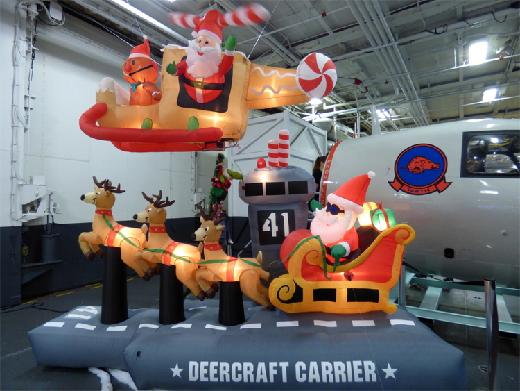 An inflatable model of an aircraft carrier with a Santa-figure landing a deer-pulled sleigh on it, with another Santa flying a helicopter above