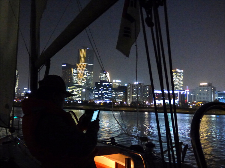 Silhouette of a person on a sailboat with lit-up city buildings on the riverbank in the distance