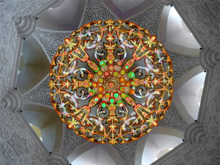 An intricate, mandala-like chandelier hanging from a white stone arched ceiling, photographed from below