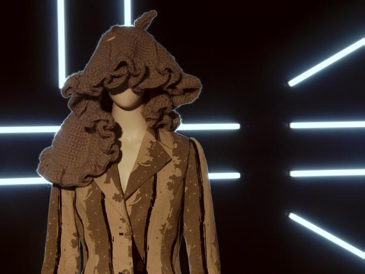 A mannequin wearing a grey coat and a shapeless grey knit thing on its head with fluorescent tubes forming a geometric pattern in the background