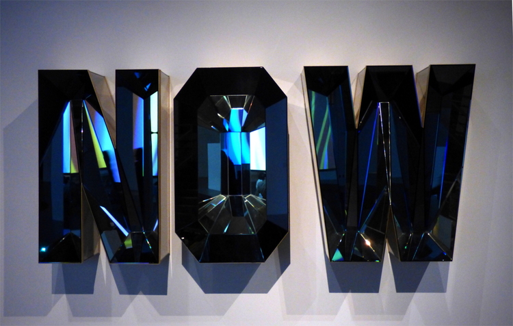 An artwork consisting of the letters 'Now' built out of mirrors and glass, reflecting light from the surrounding exhibition