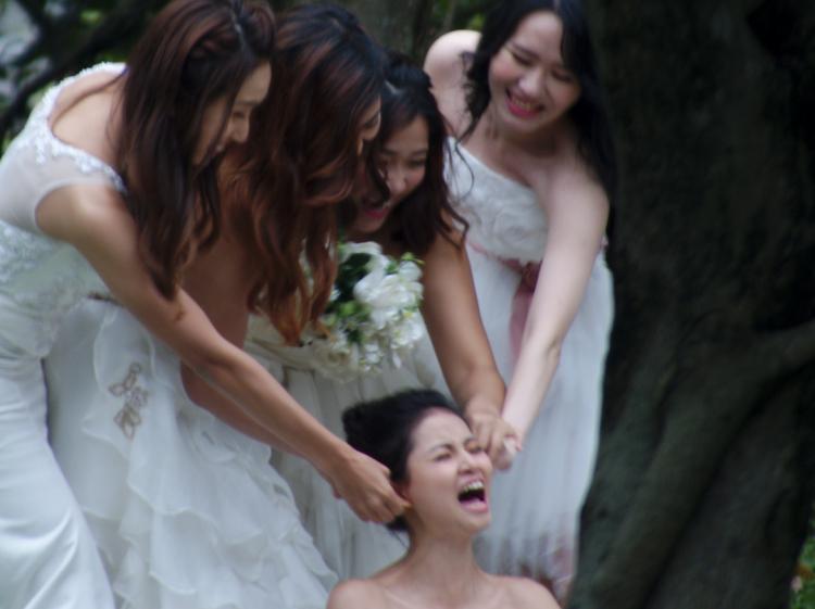 Four women in wedding dresses, one holding a bouquet, pulling on the ears of a fifth woman sitting in front of them, her face distorted in apparent pain
