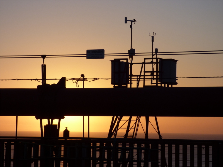 Silhouette of railings, cables, and weather measuring equipment on a bridge construction against an orange sunset sky 