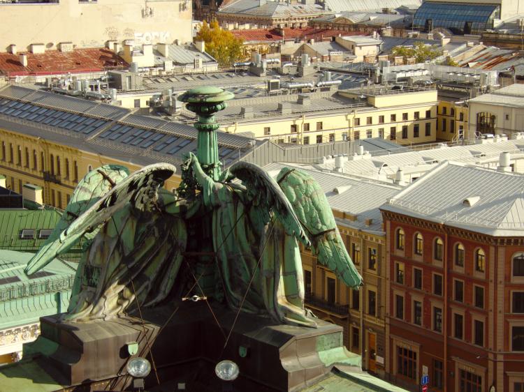 Copper sculptures depicting angels on a rooftop viewed from behind, showing pastel-coloured buildings with metal roofs in the background